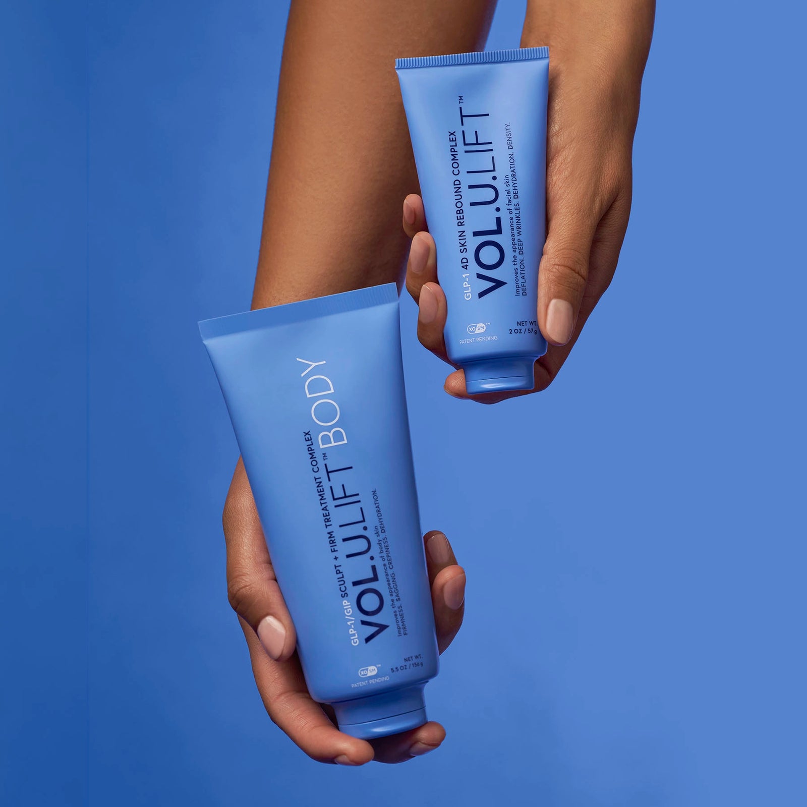 Two hands holding blue Volulift Body hand cream tubes against a solid blue background.