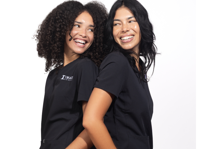 Two smiling female estheticians wearing black scrubs standing close together against a plain white background. 