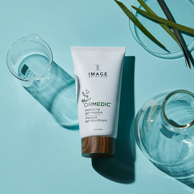 gel masque tube laying down on blue background surrounded by beaker and petri dish with plant leaves