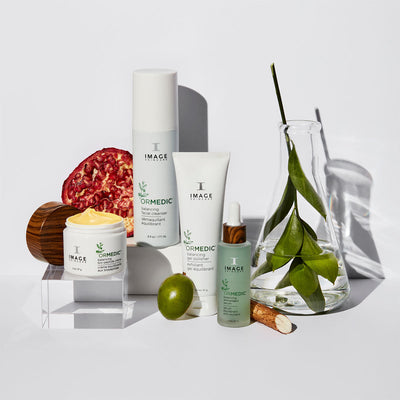 ORMEDIC product collection on white background with sliced pomegranate and plant leaves in beaker